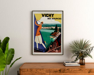 Vintagematkajuliste – Vichy, lähteet ja matkailu, kehystetty puulaatikon yläpuolelle, kasvien välissä.