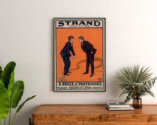 Vintage-juliste A Brace of Partridges, Strand Theatre, kehystettynä puupöydän yläpuolella kasvien välissä.