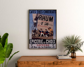 Vintage-juliste Épicerie Lefèvre, kahvit ja suklaat, kehystettynä puupöydän yläpuolella kasvien välissä.