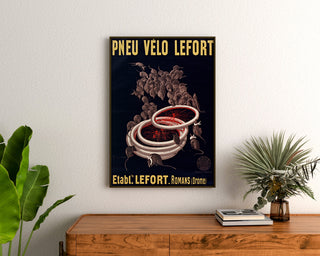 Vintage-juliste Lefort polkupyörän renkaat, kehystettynä puupöydän yläpuolella kasvien välissä.